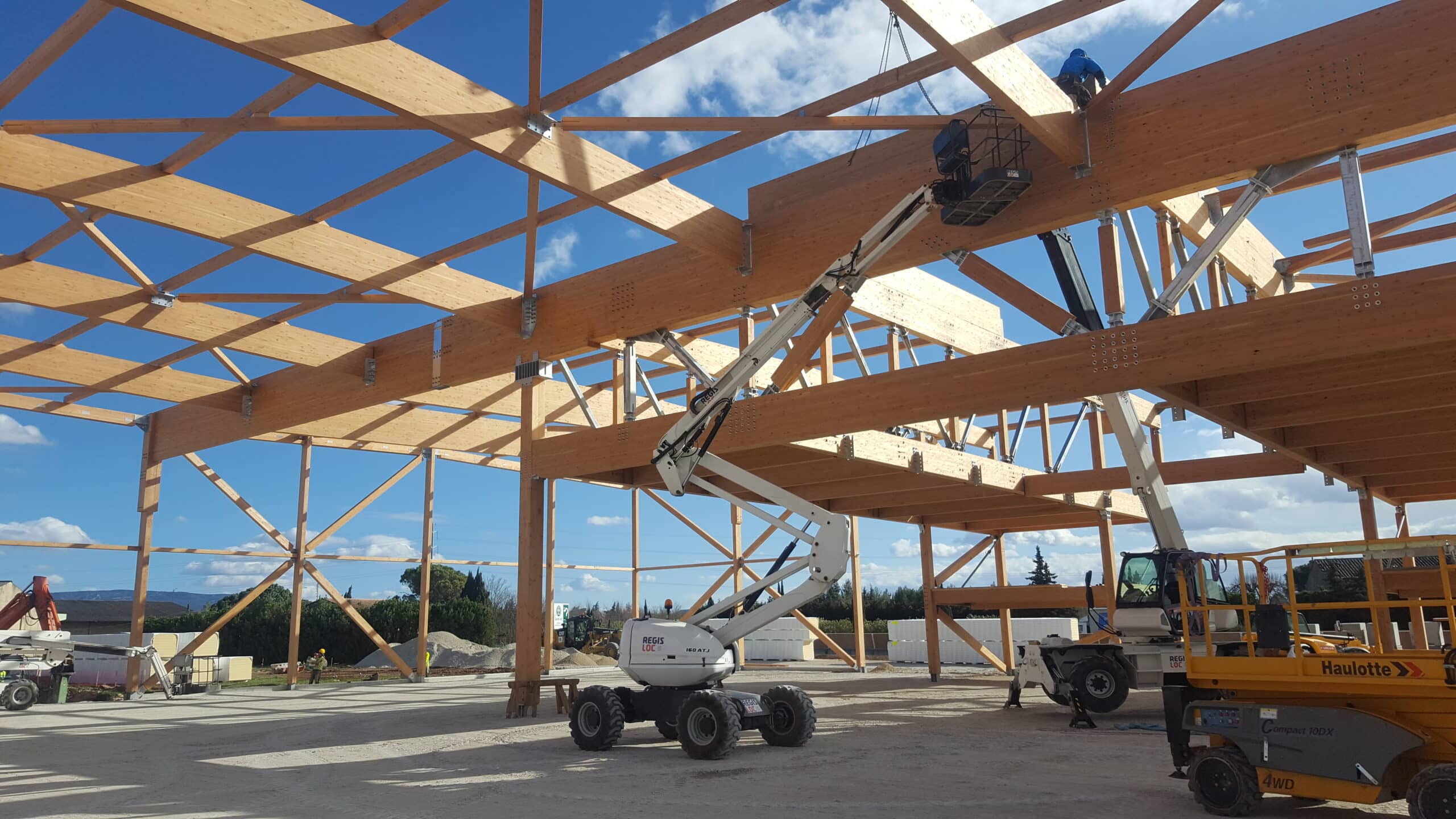 Montage de charpente en bois lamellé-collé sur chantier, ouvriers en nacelles, structure bois MONOLOG sous ciel bleu